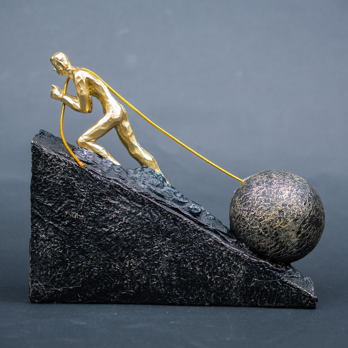 A decorative sculpture of a gold figure pushing a large metallic ball up a dark textured triangular incline, photographed against a smooth dark gray studio background.