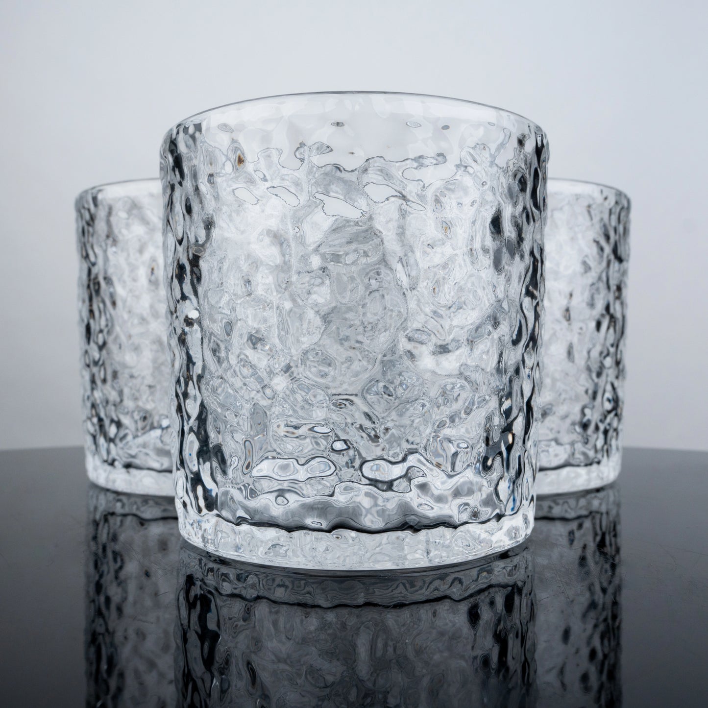 Three textured glass tumblers on a reflective surface with a light gray background