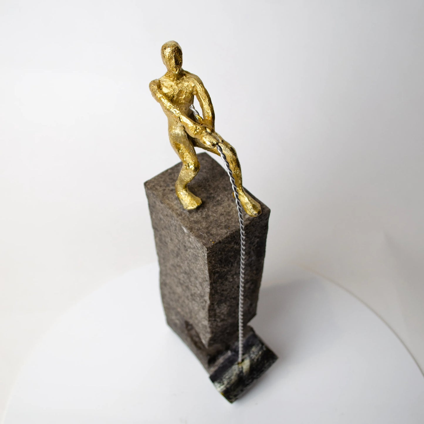 Gold pulling figure sculpture on textured charcoal stone base with hanging weight.