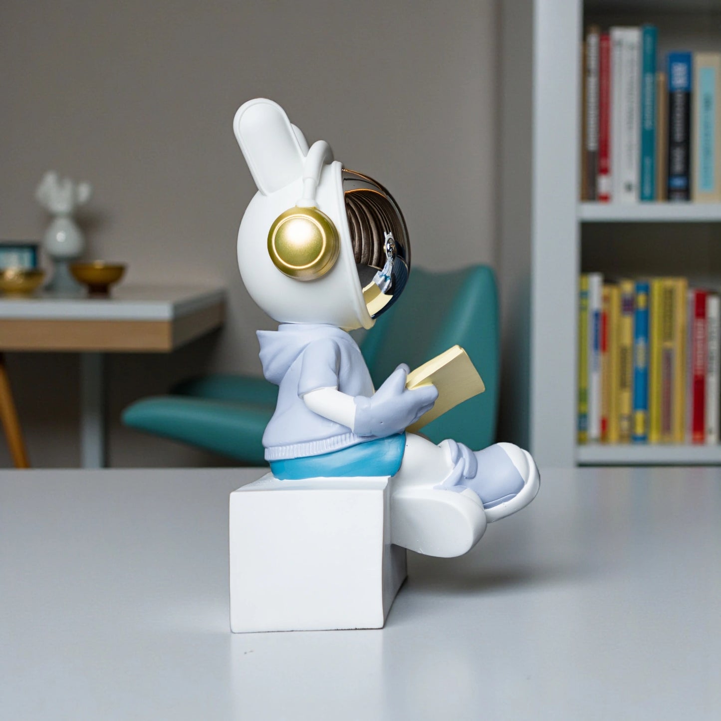 Astronaut figurine with chrome visor sitting cross-legged on a block while reading a book, wearing a white suit, gold accents, and blue pants; modern resin décor showpiece