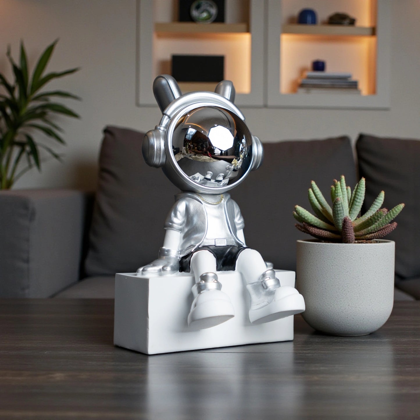 Silver astronaut figurine with chrome visor sitting casually on a white block, wearing metallic silver suit and black shorts; modern resin décor showpiece