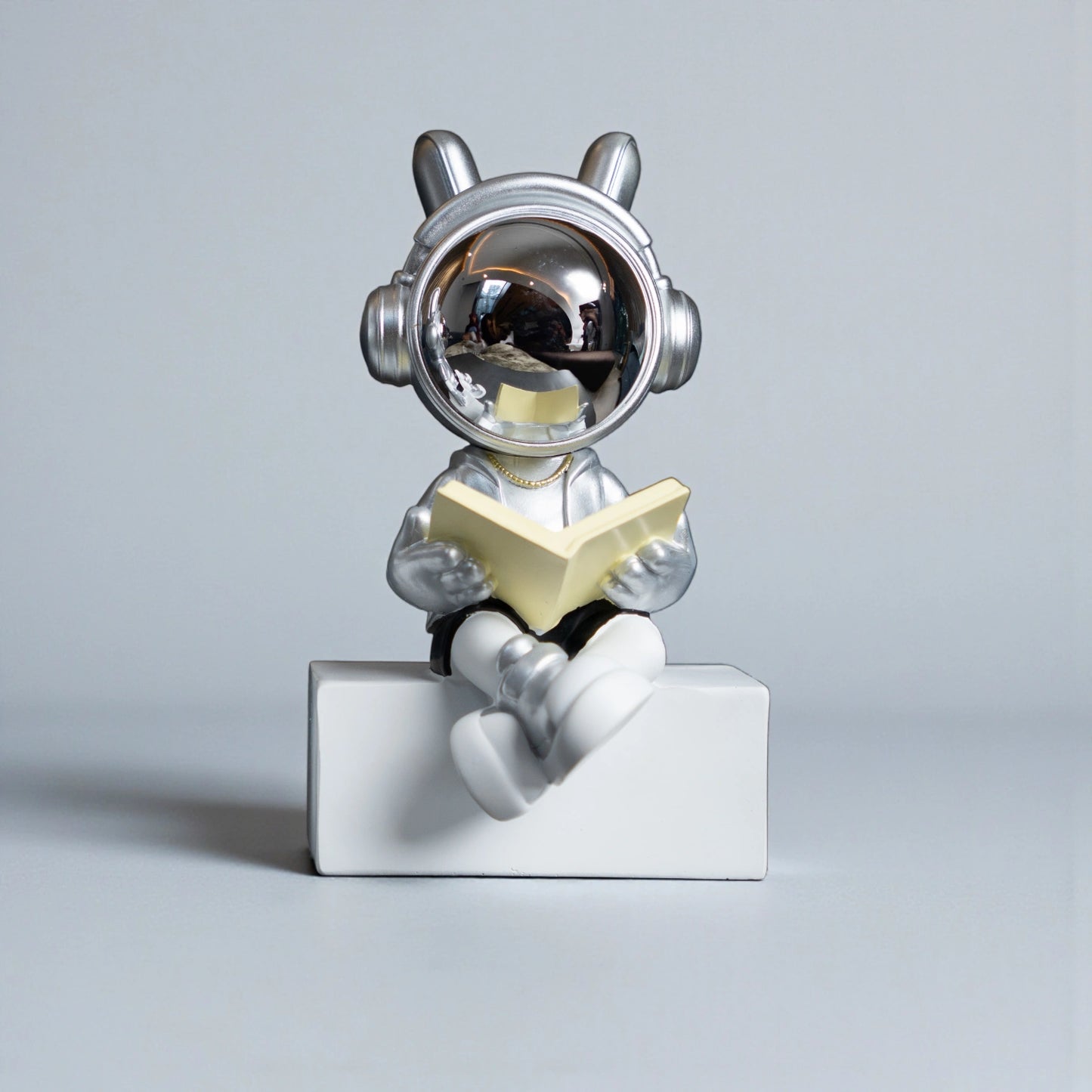 Astronaut figurine with chrome visor, silver suit, black shorts and cross-legged reading pose sitting on a white block; modern resin décor showpiece