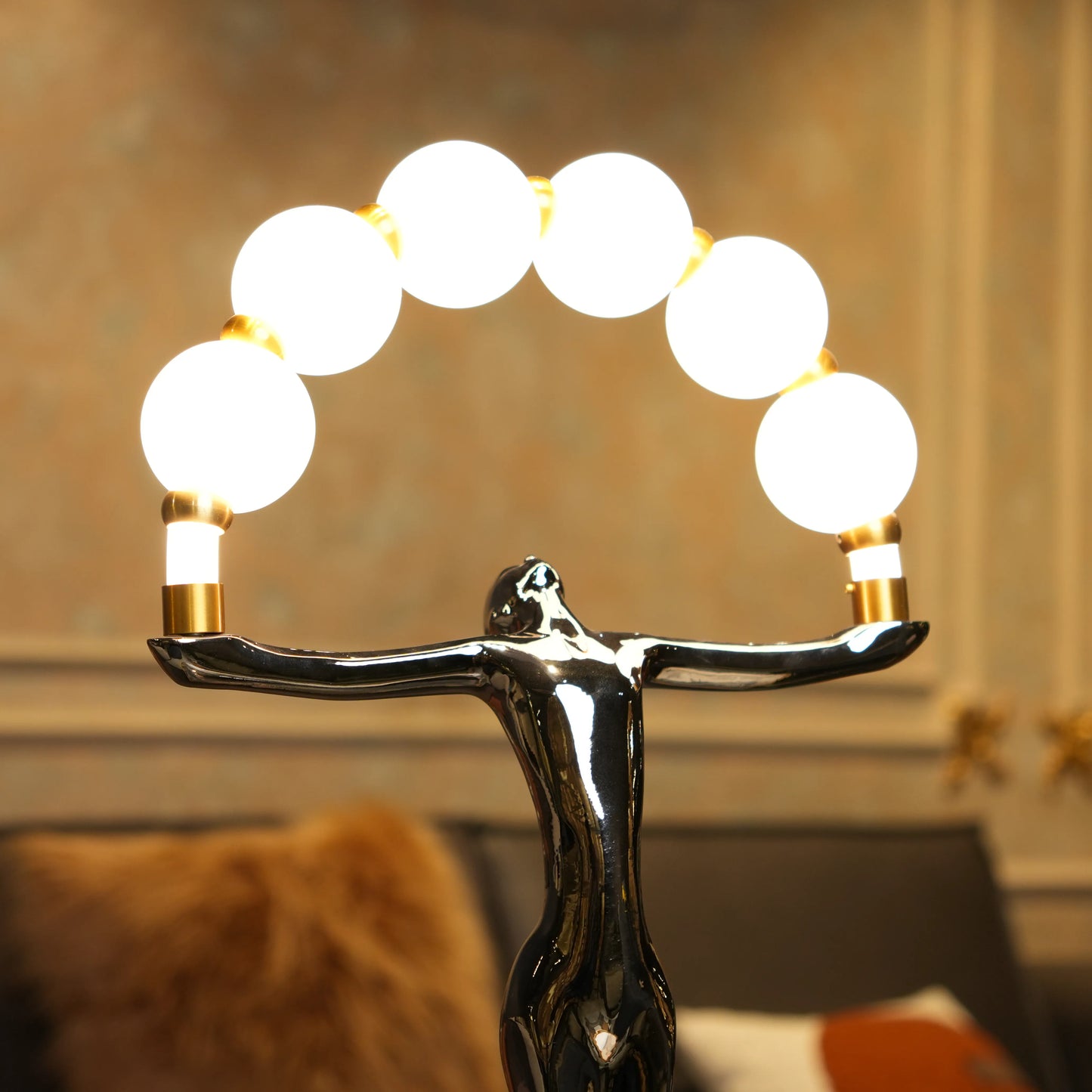 Small electroplated black human-form sculpture holding an arc of glowing warm orb lights with gold accents on a black circular base.