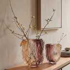 Two textured glass vases in amber and dusty rose tones, filled with delicate flowering branches, styled on a wooden console table against a warm beige wall.