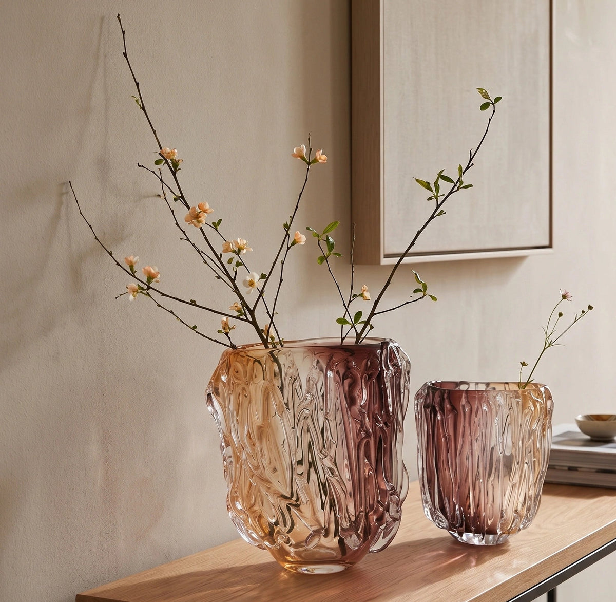 Two textured glass vases in amber and dusty rose tones, one large and one small, styled on a light wood console table with branches and books in a warm, minimalist interior.
