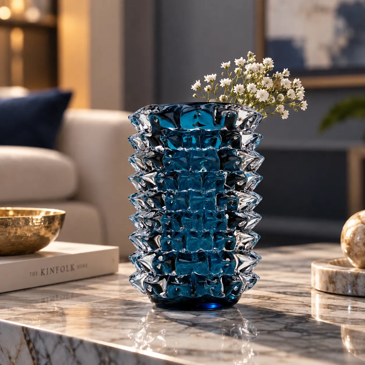 A textured, faceted cobblestone-style glass vase in a deep blue color, filled with small white baby's breath flowers, set on a polished marble table in a modern, stylishly decorated living room.