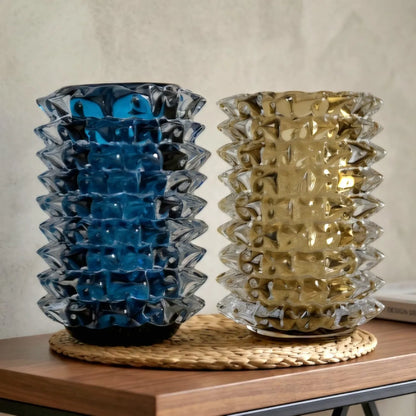 Two textured, stacked glass vases with a wavy, accordion-like pattern, one in translucent blue and one in translucent gold, displayed on a woven placemat on a wooden table against a neutral wall.