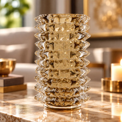 A clear textured glass vase with a geometric, faceted pattern and a fluted base, displayed on a polished marble table in a warm, softly blurred living room setting.