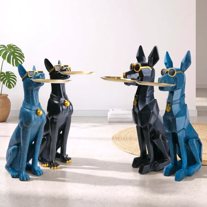 Four stylized dog statues arranged in two pairs facing each other, holding gold trays. Two statues are glossy blue and two are glossy black, all wearing gold goggles and collars.