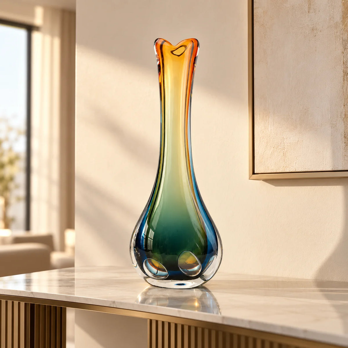 A tall, sculptural glass vase with a gradient color transition from amber orange at the top to deep teal and blue at the base, displayed on a marble console table in a modern, sunlit room.