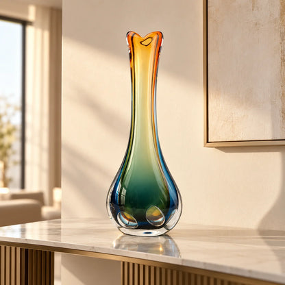 A tall, sculptural glass vase with a gradient color transition from amber orange at the top to deep teal and blue at the base, displayed on a marble console table in a modern, sunlit room.