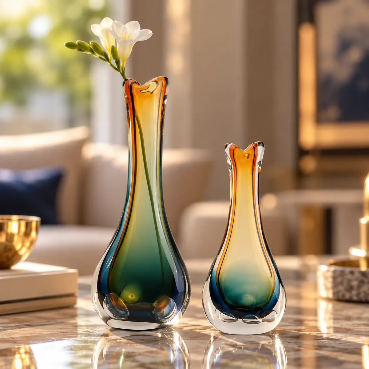Two gradient glass bud vases with amber, teal, and green color transitions, displayed on a polished marble table in a bright, modern living room setting.