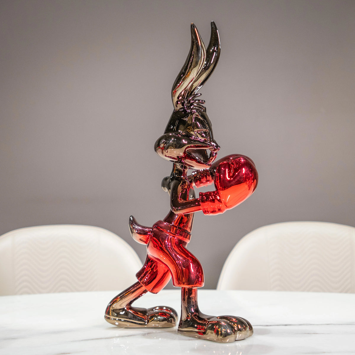 A glossy, metallic red and black cartoon rabbit figurine standing on a white marble table with blurred white chairs in the background.