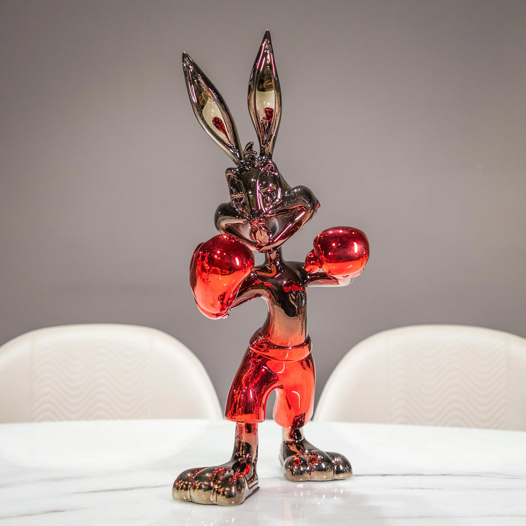 A glossy, metallic red and black cartoon rabbit figurine standing in a boxing pose on a white table, with blurred white chairs and a gray wall in the background.