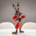 A glossy, metallic red and black cartoon rabbit figurine standing in a boxing pose on a white table, with blurred white chairs and a gray wall in the background.