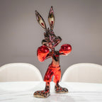 A glossy, metallic red and black cartoon rabbit figurine standing in a boxing pose on a white table, with blurred white chairs and a gray wall in the background.
