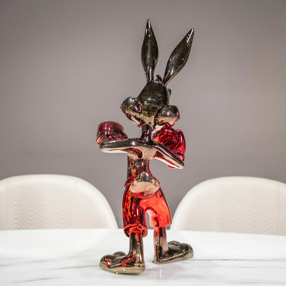 A glossy, metallic rabbit figurine with red accents, standing on a white marble table with two white chairs in the background.