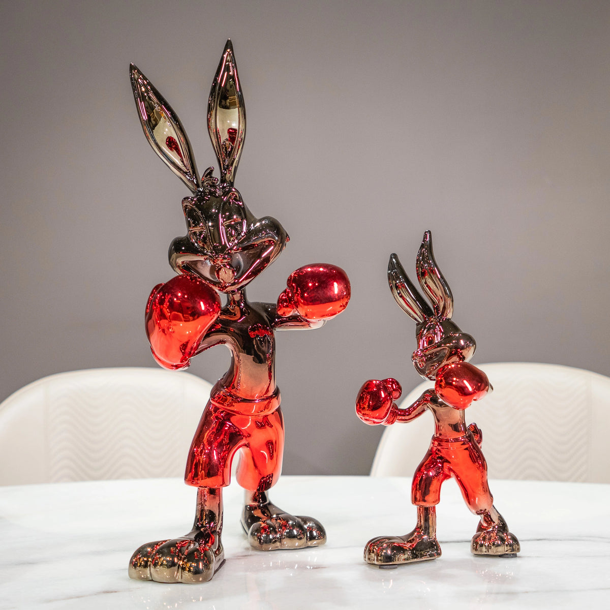 Two glossy red and metallic rabbit figurines posed as boxing statues on a white marble-like surface, with blurred white chairs and a gray wall in the background.