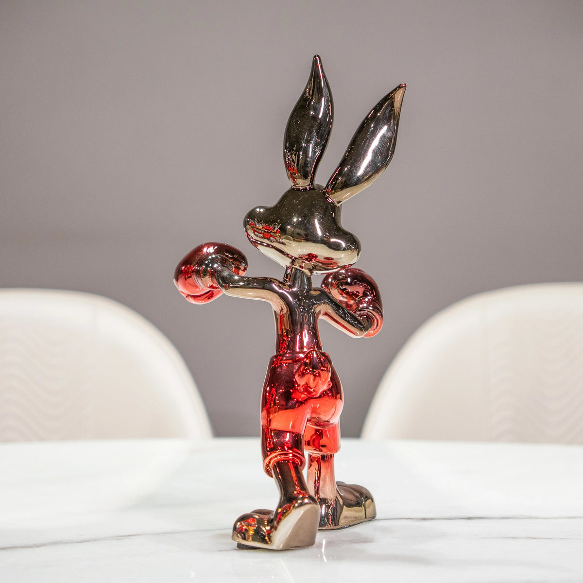 A glossy, metallic cartoon rabbit figurine standing on a white table, with two blurred white chairs against a neutral gray background.