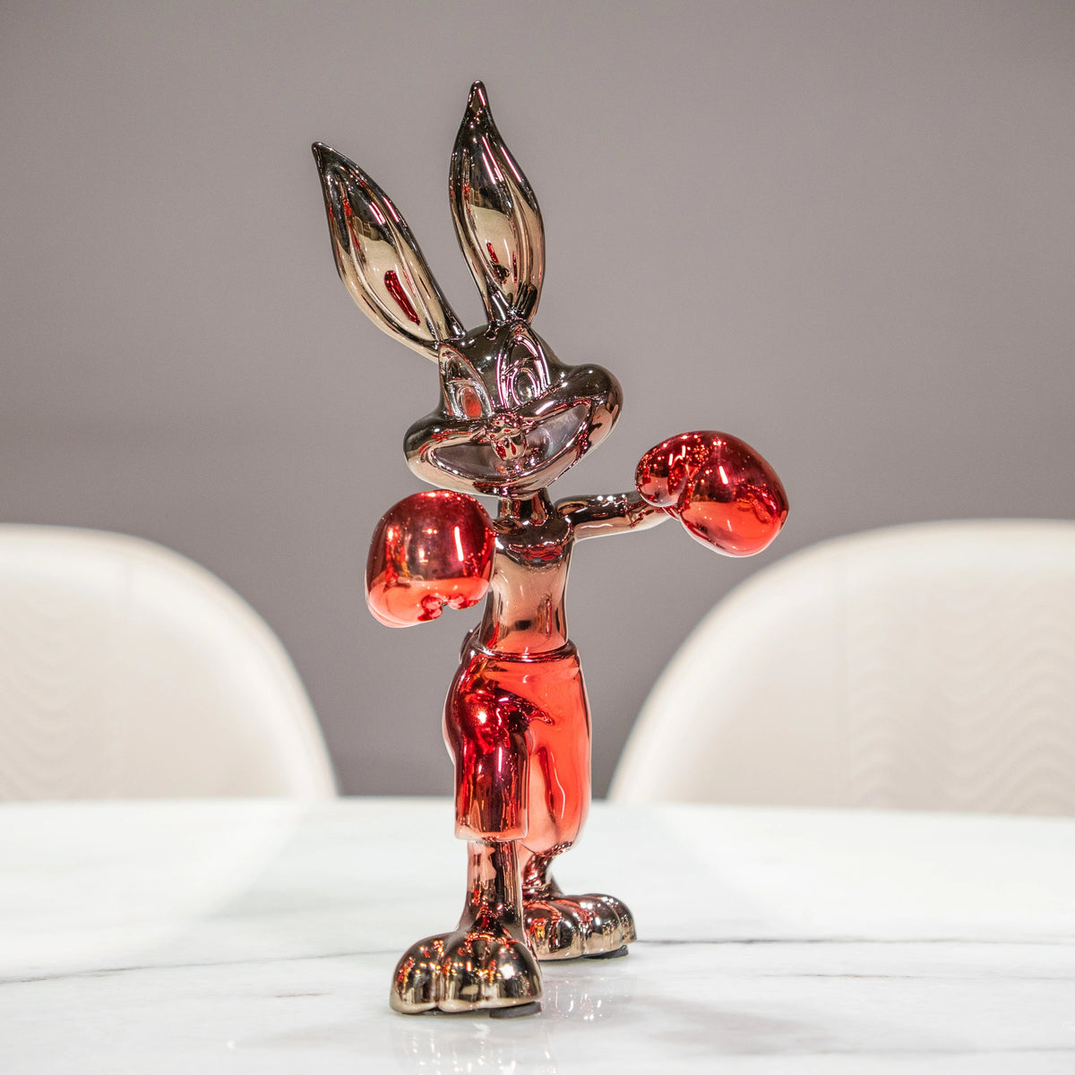 A glossy, metallic silver cartoon rabbit figurine wearing red boxing gloves and red shorts, standing on a white marble table with blurred white chairs in the background.