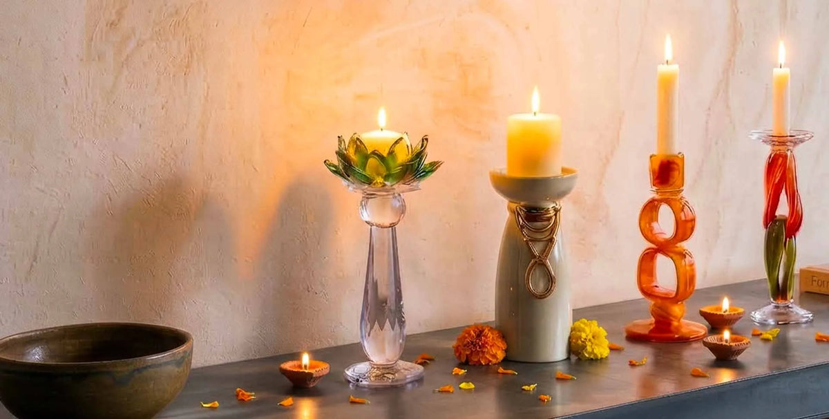 A decorative arrangement of lit candles and small oil lamps on a tabletop against a warm textured wall. The set includes a clear glass pedestal holder with a green lotus-shaped candle, a cream ceramic holder with a yellow candle, an orange sculptural candle holder, and other small candle holders, with flower petals scattered around.