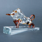 A transparent glass bull figurine with amber-brown accents on its horns, legs, and tail, standing on a clear rectangular glass base