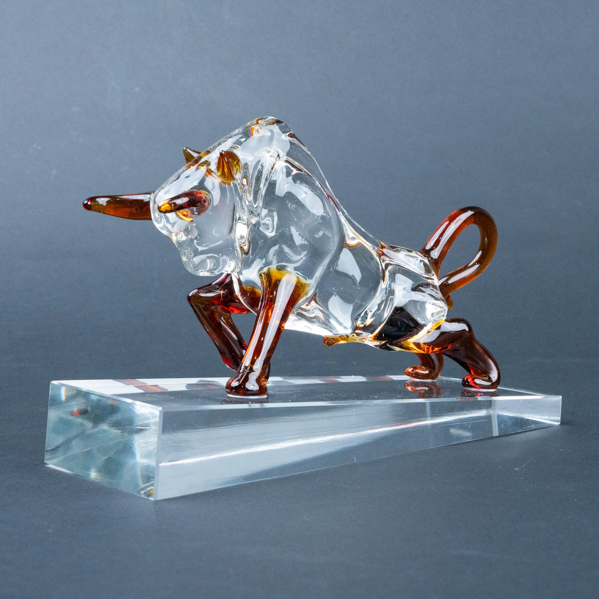 A transparent glass bull figurine with amber-brown accents on its horns, legs, and tail, standing on a clear rectangular glass base