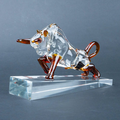 A transparent glass bull figurine with amber-brown accents on its horns, legs, and tail, standing on a clear rectangular glass base