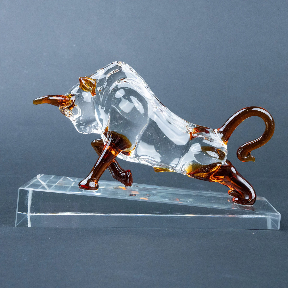 A transparent glass bull figurine with amber-brown accents on its horns, legs, and tail, mounted on a clear rectangular glass base 