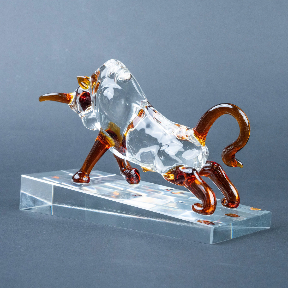A transparent glass bull figurine with amber-brown accents, posed on a clear rectangular acrylic base
