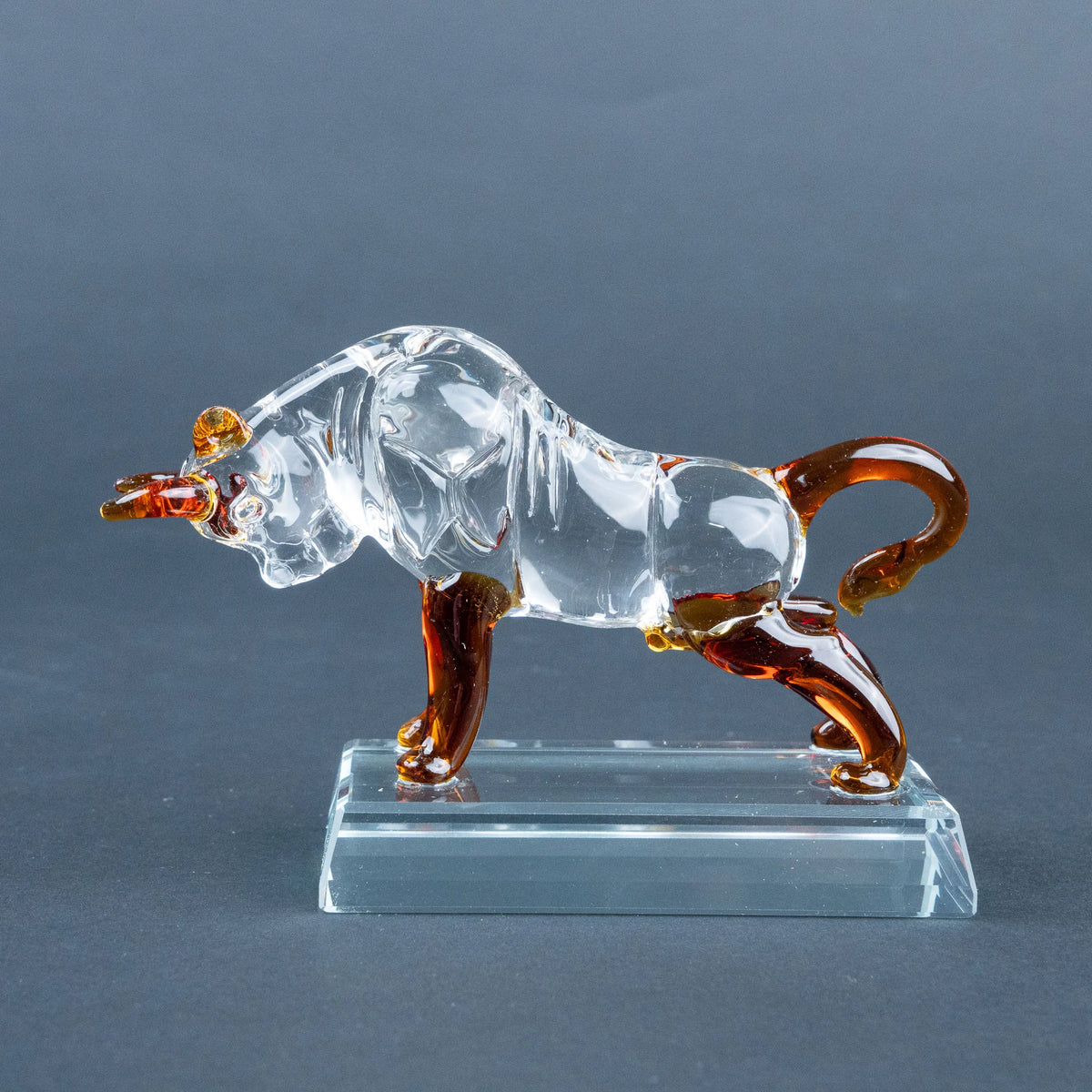 A clear glass bull figurine with amber-brown accents on its horns, legs, and tail, standing on a transparent rectangular glass base