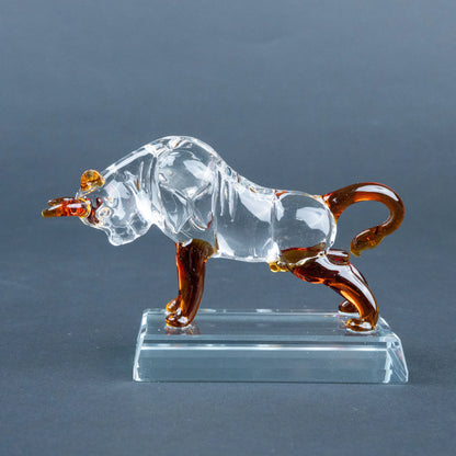 A clear glass bull figurine with amber-brown accents on its horns, legs, and tail, standing on a transparent rectangular glass base