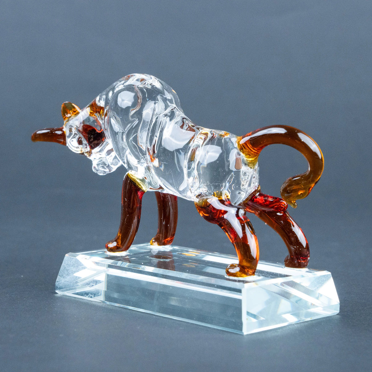 A clear glass bull figurine with amber-brown legs, tail, and horns, standing on a transparent rectangular glass base.