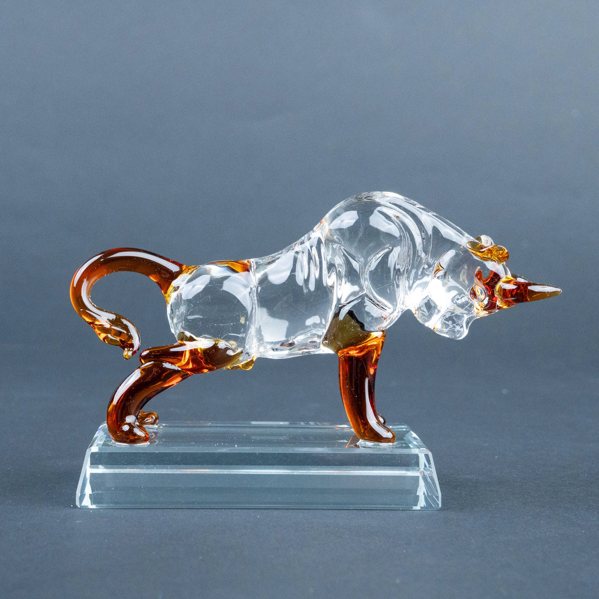 A clear glass bull figurine with amber-brown accents on its horns, legs, and tail, standing on a transparent rectangular glass base