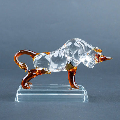 A clear glass bull figurine with amber-brown accents on its horns, legs, and tail, standing on a transparent rectangular glass base