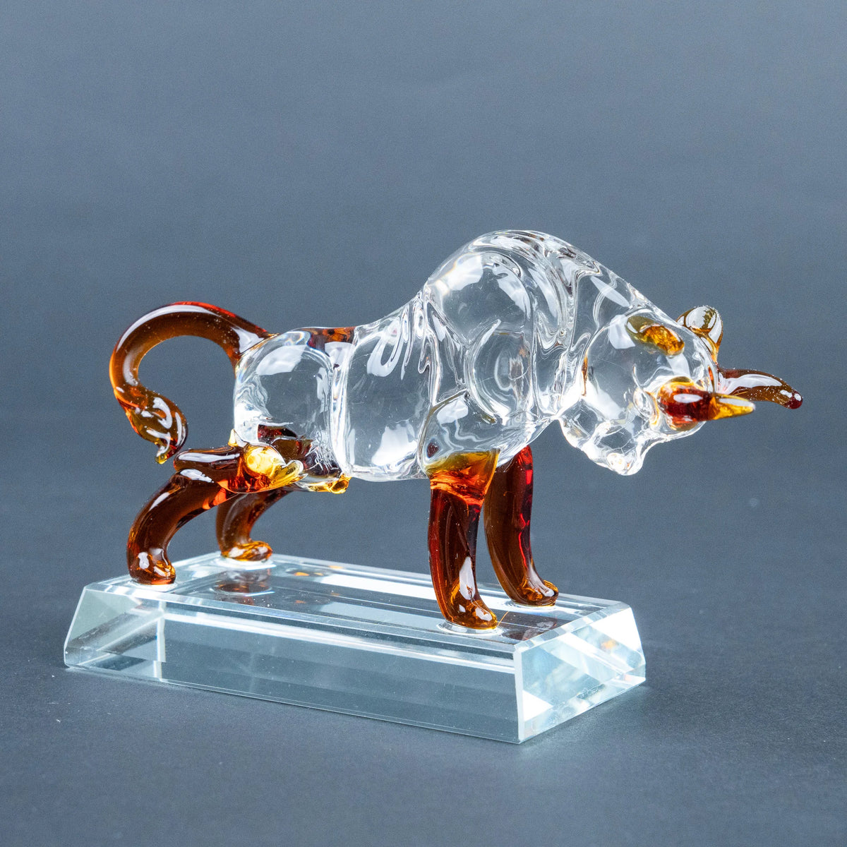 A bull figurine made of transparent clear glass with amber-brown accents on its legs, horns, and tail, standing on a clear rectangular glass base 