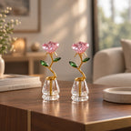 Two crystal flower vase ornaments with pink faceted rose petals, green crystal leaves, and gold stems, displayed on a wooden table in a softly lit living room.