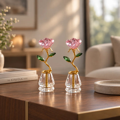 Two crystal flower vase ornaments with pink faceted rose petals, green crystal leaves, and gold stems, displayed on a wooden table in a softly lit living room.