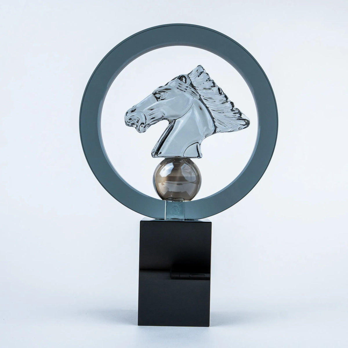 A modern glass sculpture featuring a polished silver horse head inside a large circular frame, mounted on a smoky brown glass sphere and a glossy black rectangular base.