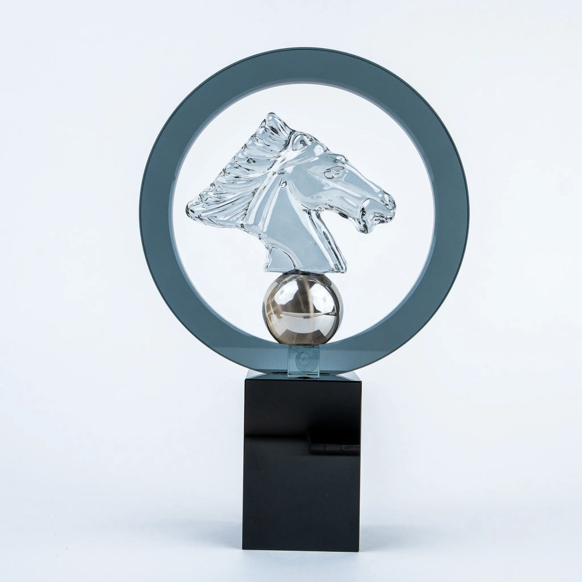 A modern glass sculpture award trophy featuring a clear glass horse head profile inside a circular frame, mounted on a polished silver sphere and a glossy black rectangular base.