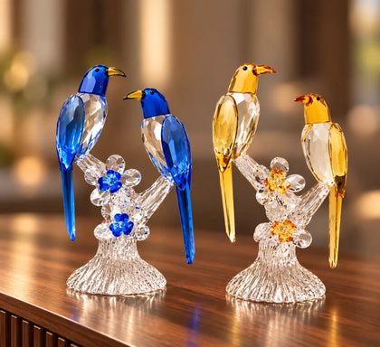 Three faceted crystal bird figurines perched on clear crystal branches with flower details, shown against a dark gray studio background. Two birds are blue, and one is yellow-gold.