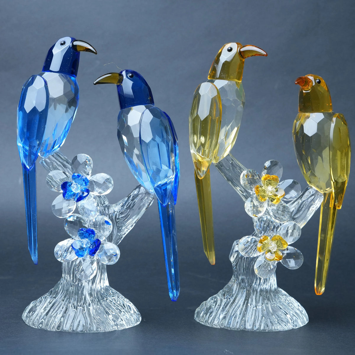 Three faceted crystal bird figurines perched on clear crystal branches with flower details, shown against a dark gray studio background. Two birds are blue, and one is yellow-gold.