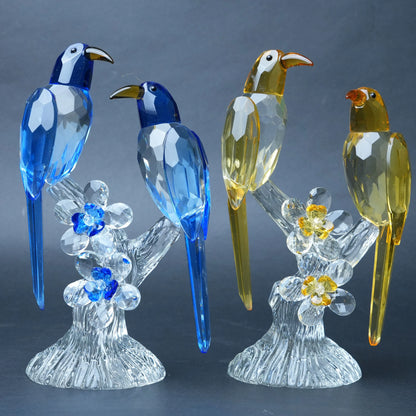 Three faceted crystal bird figurines perched on clear crystal branches with flower details, shown against a dark gray studio background. Two birds are blue, and one is yellow-gold.