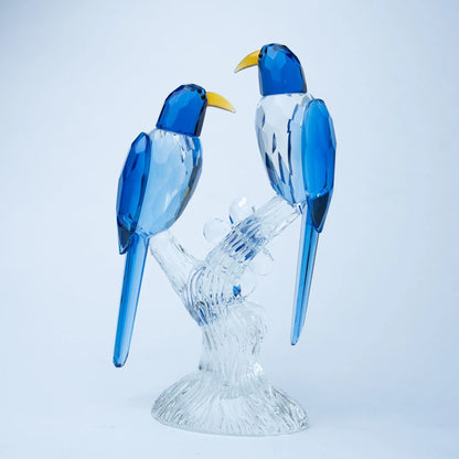 A decorative figurine of two blue birds perched on a clear glass branch and base, photographed