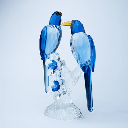 A decorative glass figurine of two blue birds perched on a clear, faceted branch and base,