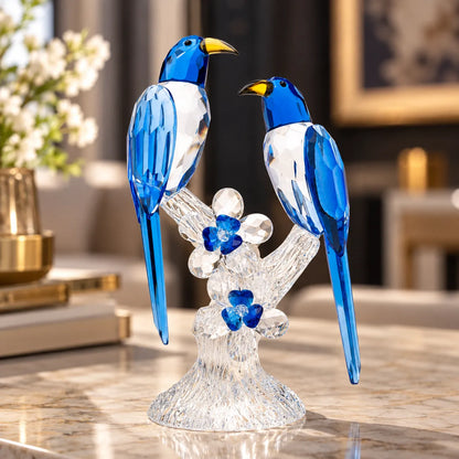A decorative crystal figurine of two blue-and-white birds perched on a clear floral branch with blue flower accents, displayed on a polished marble surface in a softly blurred indoor setting.