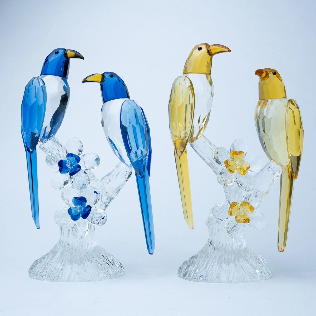 Two decorative bird figurines on clear crystal branches against a light gray background. The left pair is blue and white, and the right pair is yellow and white.