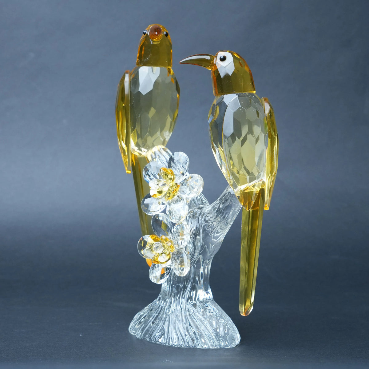 A decorative crystal bird figurine featuring two faceted amber-yellow birds perched on a clear transparent branch with small flower details