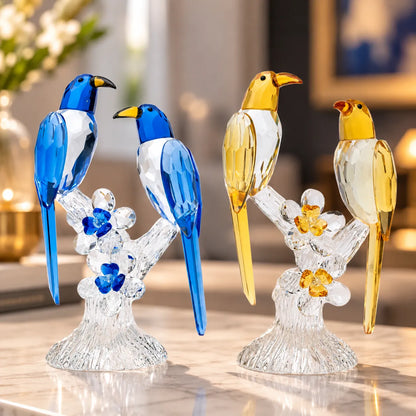 Two decorative bird figurines on clear crystal branches, shown in blue-and-white and yellow-and-white colorways on a polished marble surface with a softly blurred indoor background.
