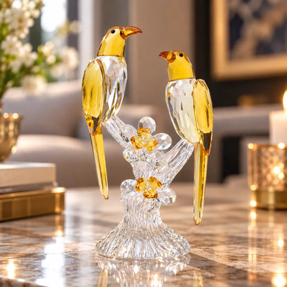 A decorative crystal bird figurine featuring two clear faceted birds with translucent amber-yellow bodies and beaks, perched on a clear crystal branch with small amber flower accents, displayed on a polished marble surface in a softly lit interior setting.
